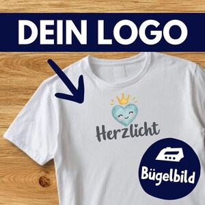 Puede incluir: Una camiseta blanca con un gráfico de un corazón azul claro con una corona amarilla y una cara sonriente. Debajo del corazón está el texto Herzlicht en gris oscuro. Una flecha azul oscuro señala el gráfico y un círculo azul oscuro contiene un icono.