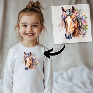 May include: A white long-sleeve shirt featuring a watercolor illustration of a horse's head adorned with a floral crown. The horse has brown and tan fur, with a white stripe down its face. The flowers are pink, purple, and blue.