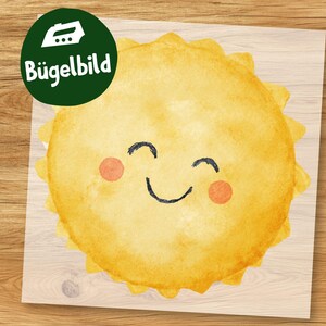 Puede incluir: Un parche termoadhesivo amarillo en forma de sol sonriente con una cara feliz, con una boca negra curvada y dos mejillas rosadas. El parche está sobre un fondo claro de vetas de madera, con un círculo verde en la esquina superior izquierda que dice "Bügelbild".