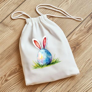 K&ouml;nnte beinhalten: Eine wei&szlig;e Baumwoll-Kordeltasche mit einer Aquarell-Illustration eines Ostereis mit Hasenohren. Das Ei ist blau, rosa und gelb mit gr&uuml;nem Gras am Boden.