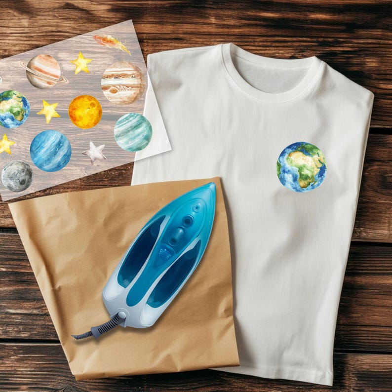 May include: A white t-shirt with a small Earth graphic, a sheet of planet and star decals, a blue and white iron, and a brown paper sheet, all on a wooden surface. The image suggests a DIY project or craft activity.