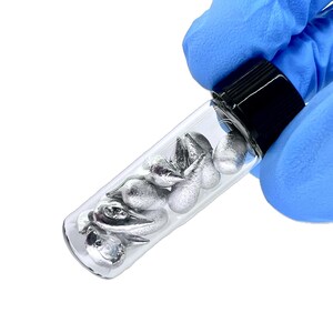 May include: A small, clear glass vial with a black screw-top lid containing several irregular, metallic silver-colored solid pieces. The vial is cylindrical in shape and holds multiple textured, teardrop-shaped metallic fragments inside.