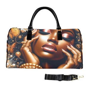 May include: A black duffel bag featuring a graphic print of a person with dark skin, closed eyes, and glossy lips, adorned with gold jewelry and surrounded by orange butterflies and pearls. The bag has black handles and a detachable black shoulder strap.