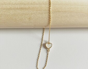 May include: A delicate gold-colored necklace featuring a small, heart-shaped pendant with a white center. The necklace is displayed against a light-colored wooden dowel and a white background, highlighting its simple elegance.