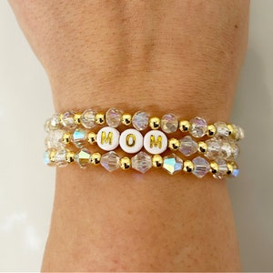 May include: A stack of three bracelets with clear iridescent beads and gold accents. The bracelets spell out the word "MOM" in white letters with gold lettering.