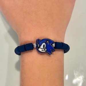 May include: A blue silicone bracelet with a silver charm featuring a cartoon image of Sonic the Hedgehog.