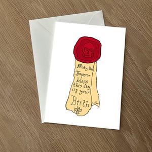 May include: A white greeting card featuring a red skull seal and a scroll with the text "May the Emperor bless this day of your Birth". The card is in a white envelope, on a wooden surface.