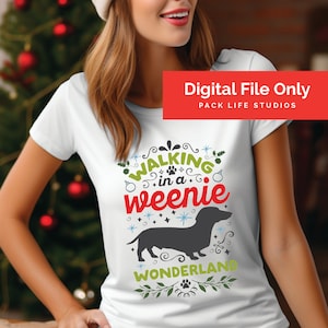 Puede incluir: Camiseta blanca con un diseño navideño con un dachshund y el texto "Walking in a weenie wonderland".