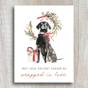 Watercolor Christmas card featuring a black dog with a red ribbon collar, sitting below a festive holiday wreath tied with a red bow, next to a red-and-white wrapped gift. The card text reads: "May your holiday season be wrapped in love."