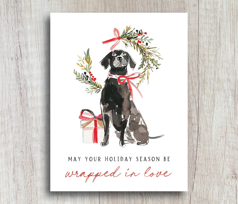 Watercolor Christmas card featuring a black dog with a red ribbon collar, sitting below a festive holiday wreath tied with a red bow, next to a red-and-white wrapped gift. The card text reads: "May your holiday season be wrapped in love."