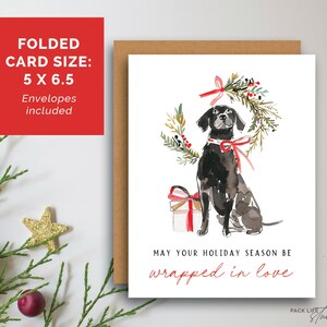 Watercolor Christmas card featuring a black dog with a red ribbon collar, sitting below a festive holiday wreath tied with a red bow, next to a red-and-white wrapped gift. The card text reads: "May your holiday season be wrapped in love."