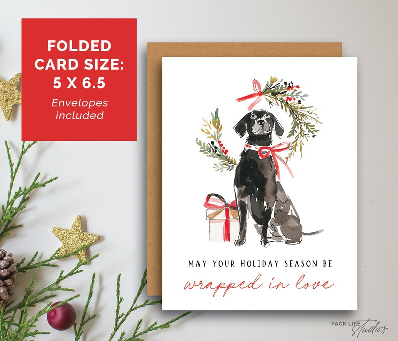 Watercolor Christmas card featuring a black dog with a red ribbon collar, sitting below a festive holiday wreath tied with a red bow, next to a red-and-white wrapped gift. The card text reads: "May your holiday season be wrapped in love."