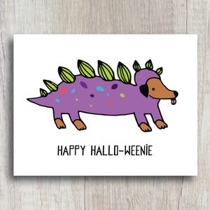 A cute and funny illustration of a dachshund dog wearing a dinosaur costume with the words happy halloweenie. Perfect greeting card for halloween and dog lovers.