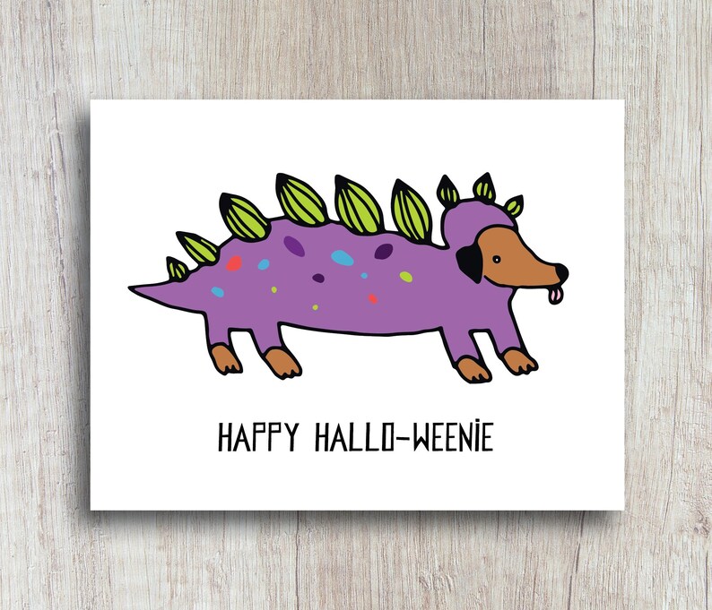 A cute and funny illustration of a dachshund dog wearing a dinosaur costume with the words happy halloweenie. Perfect greeting card for halloween and dog lovers.