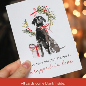 Watercolor Christmas card featuring a black dog with a red ribbon collar, sitting below a festive holiday wreath tied with a red bow, next to a red-and-white wrapped gift. The card text reads: "May your holiday season be wrapped in love."