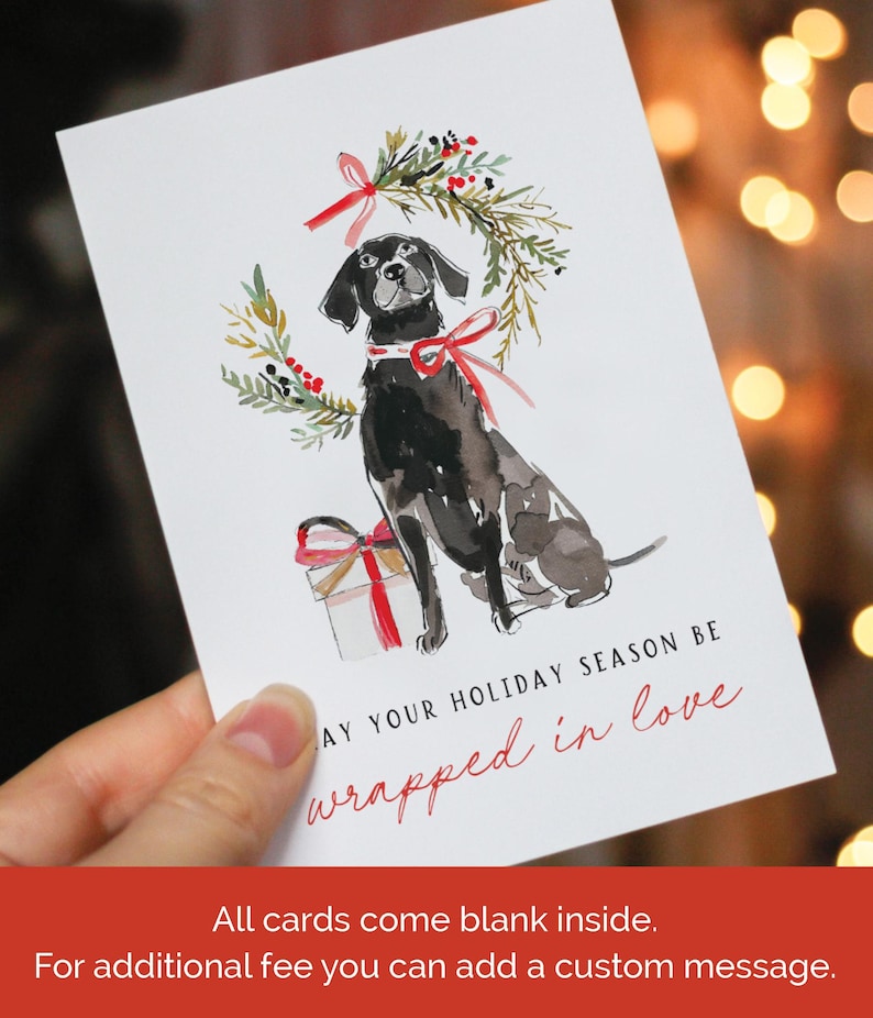 Watercolor Christmas card featuring a black dog with a red ribbon collar, sitting below a festive holiday wreath tied with a red bow, next to a red-and-white wrapped gift. The card text reads: "May your holiday season be wrapped in love."