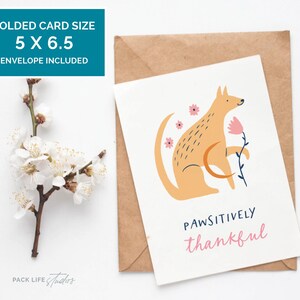 Illustrated greeting card featuring a stylized tan dog holding a pink flower, surrounded by pink flowers. Text below reads "Pawsitively Thankful" in playful navy and pink fonts.