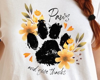 May include: White t-shirt with a black paw print surrounded by yellow flowers and green leaves. The text "Paws and give thanks" is written below the paw print.