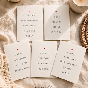 May include: Five off-white greeting cards with black sans-serif text and a small red heart icon above each message. The cards feature phrases including I hope you find something that makes you smile today, I hope today is gentle with you, I hope you laugh until your cheeks hurt, I hope you know how loved you are, and I hope something good happens to you today.