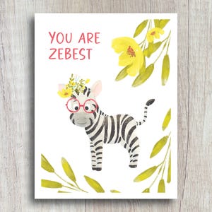 May include: A white and black zebra with a flower crown and red glasses. The zebra is standing on a white background with yellow flowers and green leaves. The text "You are Zebest" is written in pink letters at the top of the card.