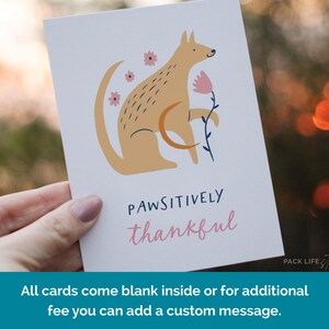 Illustrated greeting card featuring a stylized tan dog holding a pink flower, surrounded by pink flowers. Text below reads "Pawsitively Thankful" in playful navy and pink fonts.