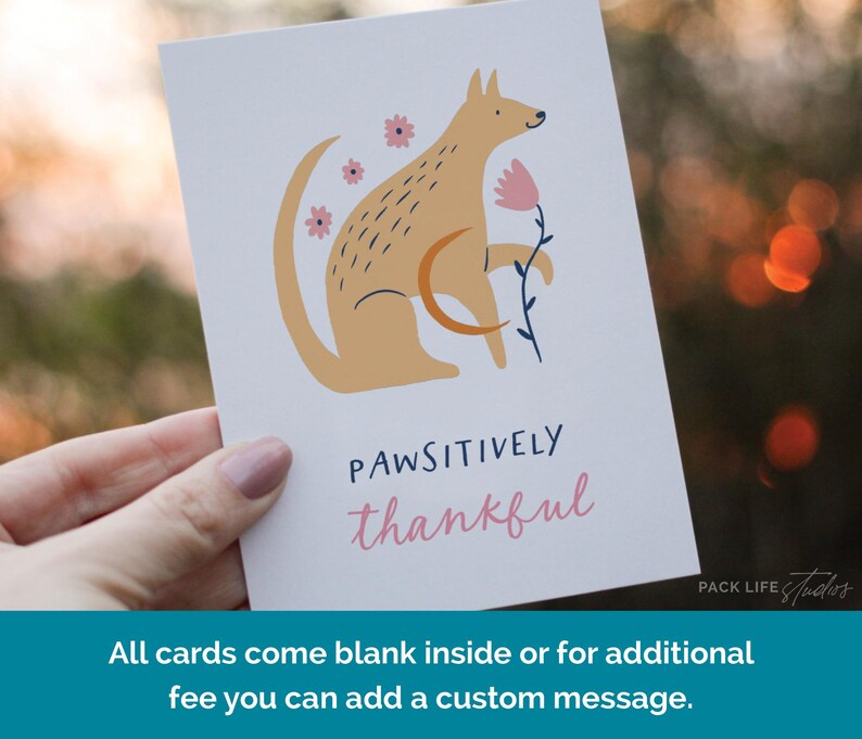 Illustrated greeting card featuring a stylized tan dog holding a pink flower, surrounded by pink flowers. Text below reads "Pawsitively Thankful" in playful navy and pink fonts.
