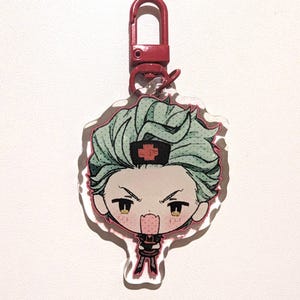May include: A clear acrylic charm featuring a cartoon character with teal hair, a red cross on their hat, and a red clasp. The character has an angry expression and is wearing a black outfit with red accents. The charm is outlined in pink.