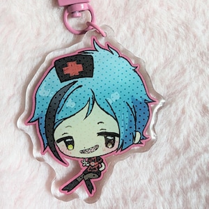 Puede incluir: Un llavero de un personaje de anime azul y rosa con una cruz roja en su sombrero. El personaje tiene el pelo azul y lleva un atuendo negro y rojo.