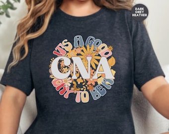 Può includere: Maglietta color grigio scuro melange con un design floreale e il testo "It's a good day to be a CNA".