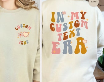 Può includere: Una felpa beige chiaro con un design grafico sul retro che dice "In My Custom Text Era" in lettere colorate in stile retrò. La parte anteriore della felpa ha un design più piccolo con le parole "Custom Text" in uno stile simile.
