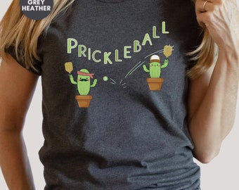 Può includere: Maglietta in jersey melange grigio scuro con una grafica di due cactus che giocano a pickleball. Il testo "Pickleball" è stampato sopra i cactus.