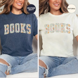 Pu&ograve; includere: Due donne indossano magliette con la parola "BOOKS" stampata in grandi lettere. Le lettere sono ricoperte da un motivo floreale. La maglietta a sinistra &egrave; blu navy melange e la maglietta a destra &egrave; naturale.