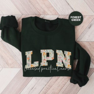 May include: A dark green sweatshirt with the letters LPN in large white letters with a floral pattern. The text "licensed practical nurse" is written below the letters.