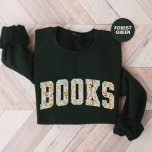 Pu&ograve; includere: Una felpa verde scuro con la parola "BOOKS" stampata sul davanti in grandi lettere bianche. Le lettere sono decorate con un motivo floreale.