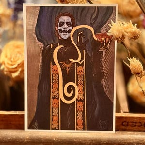 May include: A rectangular art print featuring a figure with white and black face paint, wearing a black robe with orange skull patterns and a sun emblem. A light-colored, stylized snake-like shape curves over the figure, who holds a small red cup.