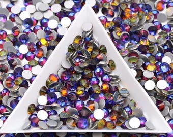 May include: A collection of iridescent rhinestones in various colors, including purple, blue, orange, and green, are scattered around a white triangular tray. The rhinestones have a flat, silver backing and are used for nail art, crafts, or jewelry making.