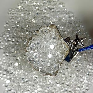 May include: A silver seashell-shaped scoop filled with clear, sparkling crystals. The scoop has a starfish and blue accents on the handle. The crystals are scattered around the scoop, creating a shimmering effect.