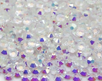 May include: A close-up view of a pile of iridescent, clear rhinestones. The rhinestones are round with a flat back and a faceted top, reflecting a rainbow of colors. They are scattered, creating a sparkling, textured surface.