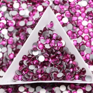 May include: Close-up of a white triangular tray filled with vibrant fuchsia rhinestones. The rhinestones are a mix of faceted gems and flat-backed, silver-backed stones. The background is filled with scattered rhinestones.