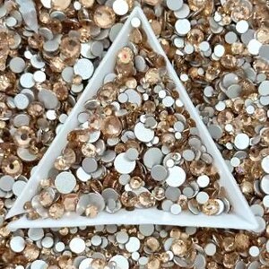 May include: A white triangular tray filled with various sizes of light brown and silver rhinestones. The tray is surrounded by a scattered assortment of similar rhinestones, creating a textured, sparkling effect. Ideal for crafting and embellishment projects.