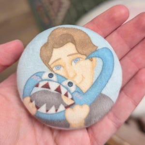 May include: A round, light blue button featuring a cartoon illustration of a person hugging a blue shark-like creature. The person has brown hair and blue eyes. The shark has a white mouth with red teeth.