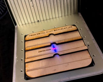 May include: A wooden jig for making 19mm metal cuff bracelets. The jig has four slots and the text "CENTER HERE" is printed on the jig. A laser pointer is shining on the jig.