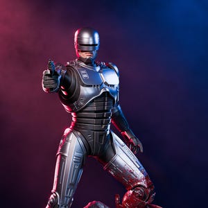 Puede incluir: Estatua detallada de RoboCop con armadura plateada y negra, sosteniendo un arma y de pie sobre una figura derrotada. La base es un diorama detallado con una escena callejera y sangre. El fondo tiene iluminación roja y azul.