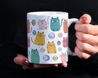 May include: White ceramic mug with a colorful pattern of cats and yarn balls. The cats are drawn in a cartoon style and are various colors, including pink, yellow, blue, and green.