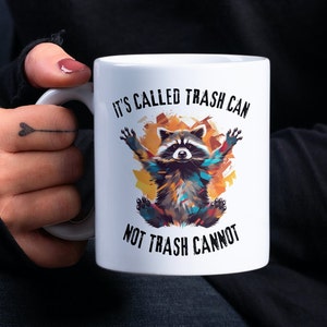 May include: White ceramic mug with a colorful raccoon illustration and the text "It's called trash can not trash cannot".
