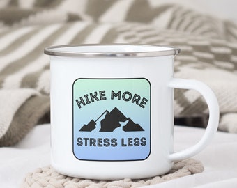 May include: White enamel camping mug with a blue, green and white gradient square design. The design features a black mountain silhouette and the text "Hike More Stress Less".