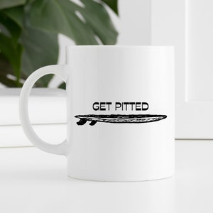 May include: White ceramic mug featuring a black surfboard illustration and the text "GET PITTED". The mug has a curved handle and is set against a white background with a green plant visible.