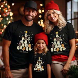 May include: A family of three wearing matching black t-shirts with a Christmas tree design featuring cats. The tree is decorated with a star and ornaments. The text "Kit's Christmas" is printed on the shirts.