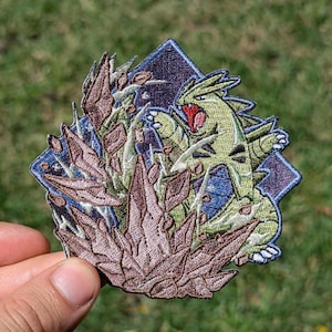 Op de afbeelding: Een geborduurde patch met een groen, tweebenig wezen met een rode mond en scherpe klauwen, geplaatst achter een cluster van bruine, grillige rotsachtige formaties tegen een donkerblauwe, ruitvormige achtergrond met een gestikte rand.