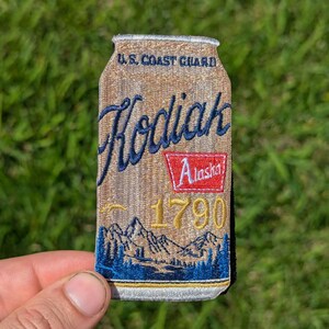May include: Embroidered patch in the shape of a beer can, featuring the text "Kodiak Alaska 1790" in blue and gold. The top reads "U.S. Coast Guard". The bottom of the patch shows a mountain and forest scene.